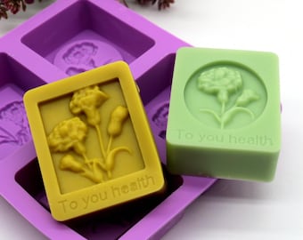May include: Two square-shaped bars of soap with a floral design and the text "To you health" on each. The soap bars are in a purple silicone mold.