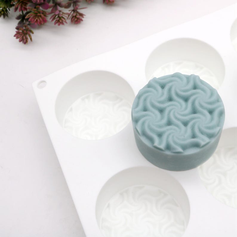 May include: A light blue, round soap with a swirling pattern sits in a white silicone mold. The mold has several circular cavities, each with a similar swirl design at the base. The soap has a textured surface.