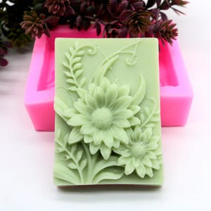 Puede incluir: Un molde de silicona rosa para hacer jabón con un diseño floral detallado. El molde presenta dos margaritas grandes y flores más pequeñas con hojas y enredaderas.