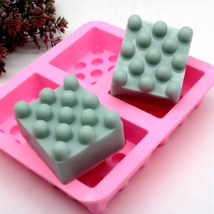 Puede incluir: Dos barras de jabón verde claro en un molde de silicona rosa. Las barras de jabón tienen una superficie texturizada con pequeños bultos. El molde tiene una forma rectangular con múltiples pequeños agujeros.