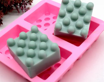 Puede incluir: Dos barras de jabón verde claro en un molde de silicona rosa. Las barras de jabón tienen una superficie texturizada con pequeños bultos. El molde tiene una forma rectangular con múltiples pequeños agujeros.