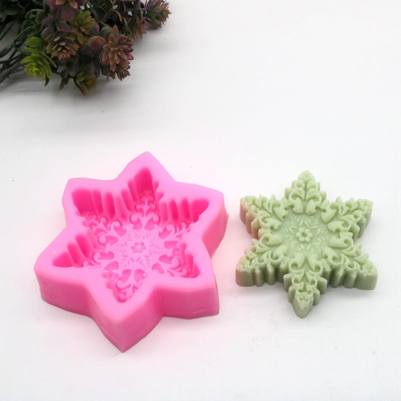 May include: Pink silicone mold shaped like a six-pointed star with a detailed snowflake design inside. A green, finished snowflake made with the mold is shown next to it.