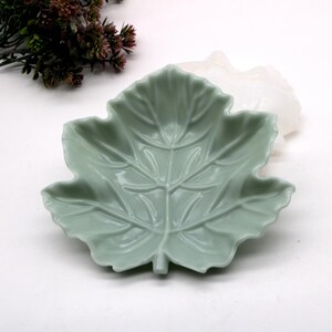 May include: A light green, leaf-shaped ceramic dish. The dish has detailed veining and a slightly raised edge. The design is a stylized maple leaf. The dish is on a white surface, with some decorative greenery in the background.