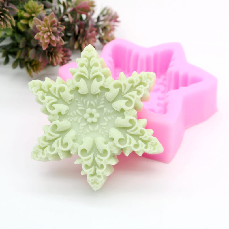 May include: A light green snowflake-shaped soap mold with a detailed design. The mold is made of silicone and is pink in color.