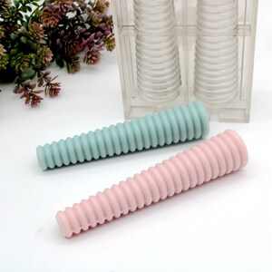 Può includere: Due candele a spirale, una blu chiaro e una rosa, sono mostrate accanto a uno stampo in plastica trasparente. Lo stampo ha due sezioni, ciascuna con una serie di anelli che creano la forma a spirale delle candele.