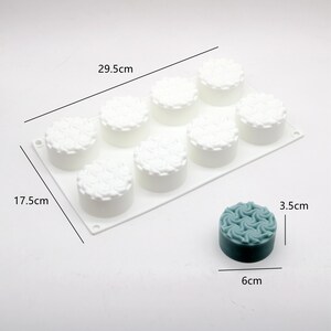 May include: A white silicone mold, 11.6 inches long and 6.9 inches wide, with eight round cavities, each with a floral design. A teal-colored, 2.4-inch diameter, 1.4-inch tall, finished product is shown next to the mold.
