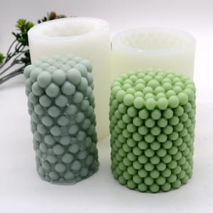 May include: Two white silicone molds for making candles. The molds are cylindrical and have a textured surface that resembles a cluster of small spheres. The molds are empty, but there are two finished candles next to them, one in each mold. The candles are green and have a textured surface that matches the molds.