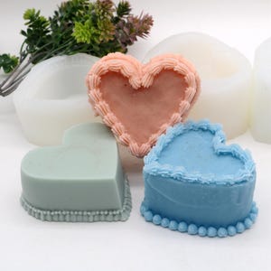 Puede incluir: Tres moldes de silicona en forma de corazón para hacer pasteles o jabones. Los moldes son de color rosa, azul y verde y tienen un borde decorativo alrededor del borde.