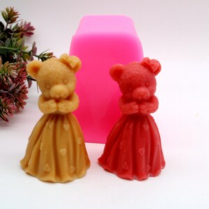 May include: Two small bear figurines wearing dresses with heart patterns. The bears are in a standing position with their hands clasped in front of them. The bear on the left is wearing a brown dress with gold hearts, and the bear on the right is wearing a red dress with red hearts. The bears are sitting on a white surface.