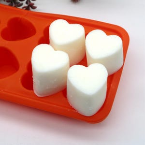 May include: Four white heart-shaped candles in a bright orange silicone mold. The candles are ready to be removed from the mold.