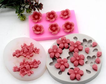 May include: Three silicone molds for making cherry blossom shaped cakes. The molds are pink, gray, and white. The molds are filled with pink cherry blossom shapes.