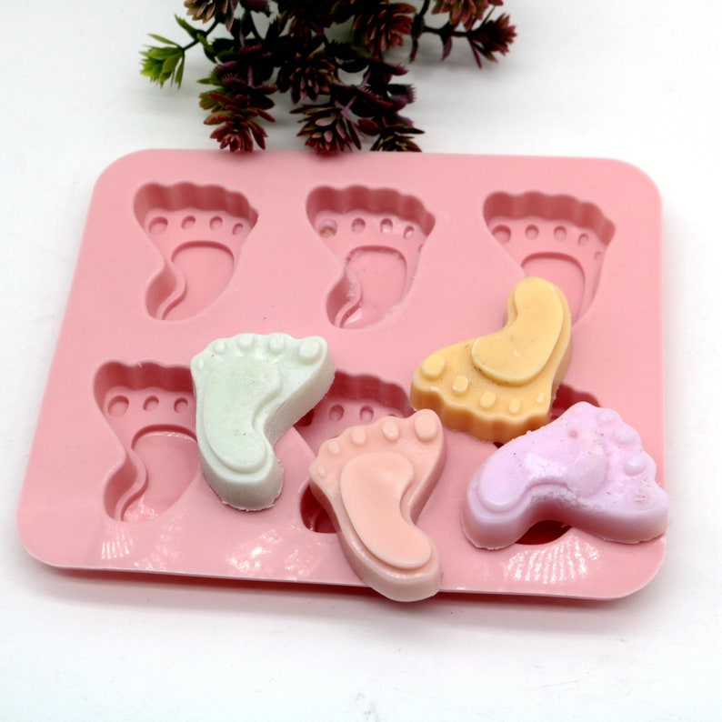 May include: Pink silicone mold with six baby footprint shapes.  Four of the shapes are filled with colorful soap in shades of pink, white, and yellow.
