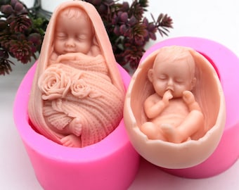 May include: Two pink silicone moulds for making baby-shaped soap or candles. One mould features a baby wrapped in a blanket with roses, the other mould features a baby in an egg-shaped shell.