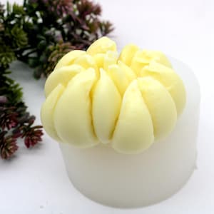 Puede incluir: Una vela cilíndrica blanca rematada con un diseño en forma de flor amarillo claro. El elemento floral tiene múltiples pétalos redondeados. El fondo es blanco con un poco de vegetación.