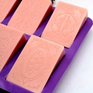 May include: A close-up of a purple silicone mold with six pink soap bars. The soap bars have intricate floral designs.