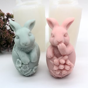 Könnte beinhalten: Zwei Hasenfiguren aus Seife, eine ist hellblau und die andere rosa. Beide sitzen in einer eiförmigen Form mit Blumendesigns.