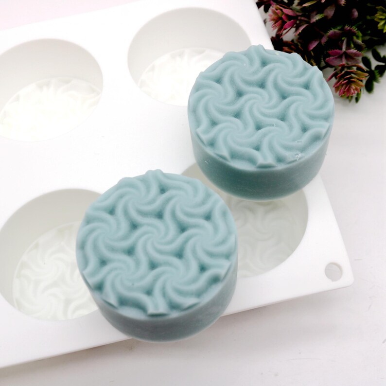 May include: Two round, light blue soaps with a swirling pattern sit in a white mold. The soaps have a textured surface and a slightly rounded shape. The mold has four circular cavities, with two soaps visible.
