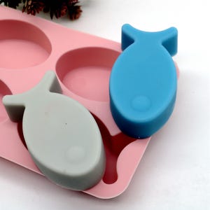 Puede incluir: Un molde de silicona rosa con dos barras de jabón con forma de pez. Una barra de jabón es gris claro y la otra es azul claro.