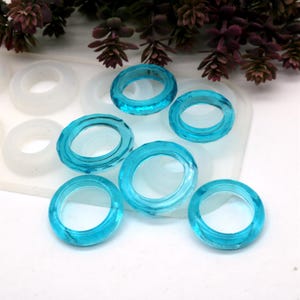 May include: Seven turquoise resin rings, each with a slightly different shape and size, are arranged on a white surface. The rings are translucent and have a glossy finish.