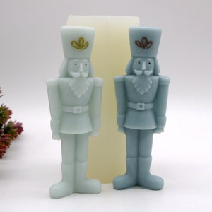 May include: Two blue nutcracker candle holders with gold accents. The nutcrackers are standing on a white surface.