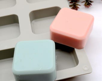 May include: Two square-shaped bars of soap, one light blue and one light pink, are sitting in a silicone mold. The mold has multiple square cavities.
