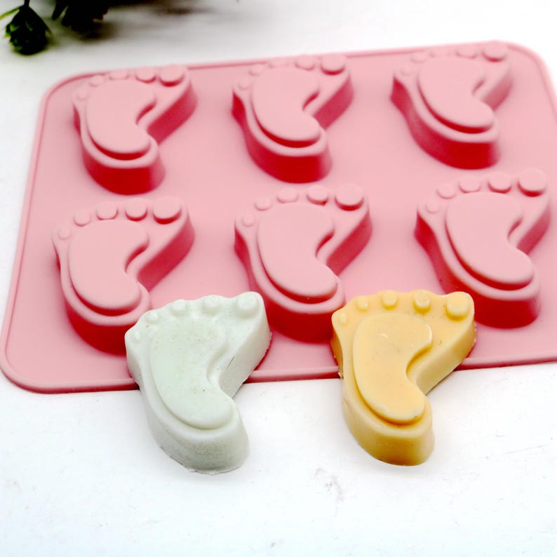 May include: Pink silicone mold with six baby footprint shapes. Two finished soap bars are shown, one white and one yellow.