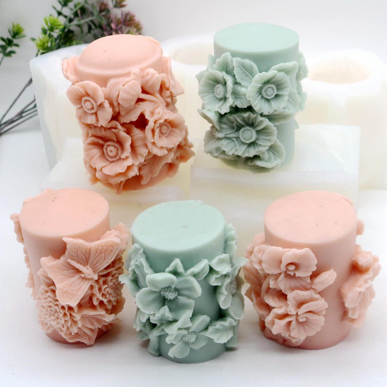May include: Five cylindrical candles with floral designs. Two candles are pink, two are green, and one is light pink. The candles are arranged in a circle on a white surface.