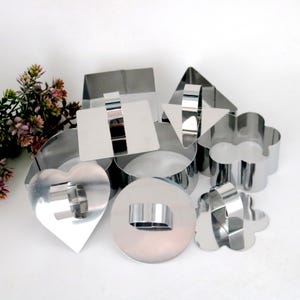 Puede incluir: Un conjunto de cortadores de galletas de acero inoxidable brillante en varias formas, incluyendo un corazón, círculo, cuadrado, triángulo y flor. Los cortadores están dispuestos sobre una superficie blanca, con un pequeño arreglo floral en el fondo.