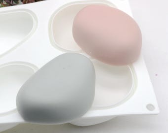 Puede incluir: Un molde de silicona blanco con cuatro cavidades con forma de piedras lisas y redondeadas. Dos de las cavidades están llenas de resina gris y rosa, que se asemejan a rocas.