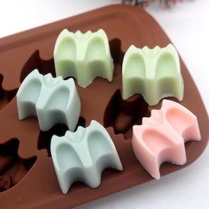 May include: A silicone mold with a brown background and five bat-shaped cavities. The cavities are filled with green, light blue, and pink wax.
