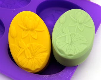 May include: Two oval-shaped silicone molds for making soap. One mold is filled with a yellow soap bar with a dragonfly and lotus flower design. The other mold is filled with a green soap bar with a dragonfly and lotus flower design.