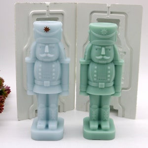 May include: Two nutcracker-shaped silicone molds, one light blue and one green, for making candles or soap. The molds are in a white plastic packaging.