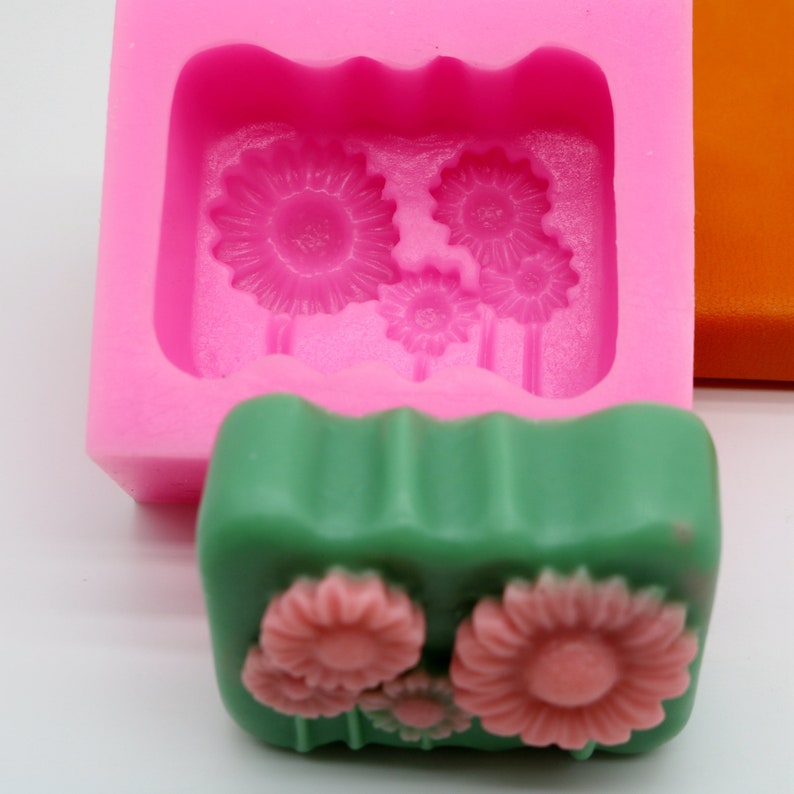 Puede incluir: Molde de silicona rosa con tres formas de flores, y una barra de jab&oacute;n verde con tres formas de flores rosas.