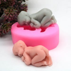 Puede incluir: Molde de silicona rosa con dos figuras de bebés. Un bebé es gris y el otro es rosa. Ambos bebés están durmiendo.