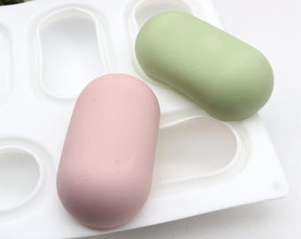 May include: A white silicone mold with six cavities shaped like elongated ovals. Two finished resin pieces are shown in the mold, one is light green and the other is light pink.
