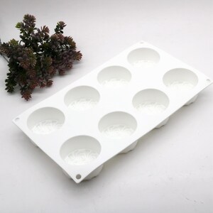 May include: A white silicone mold with eight circular cavities, each with a decorative pattern at the base. The mold is rectangular with rounded corners. A sprig of dark green and purple faux foliage is in the background.