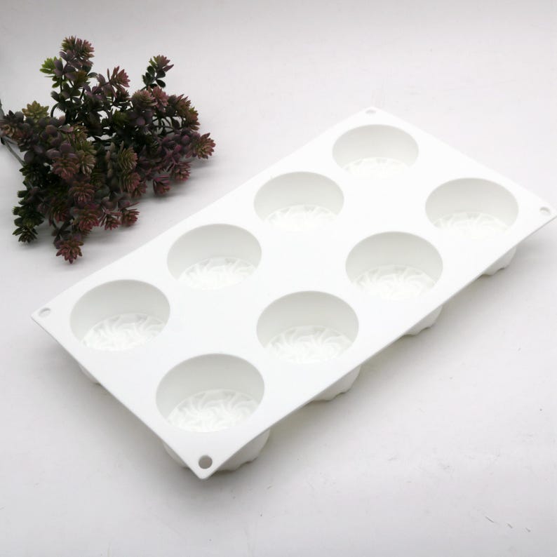 May include: A white silicone mold with eight circular cavities, each with a decorative pattern at the base. The mold is rectangular with rounded corners. A sprig of dark green and purple faux foliage is in the background.