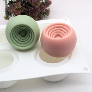 May include: A white silicone mold with six round cavities, two of which are filled with green and pink sphere-shaped soaps. The soaps have a concentric circle design.