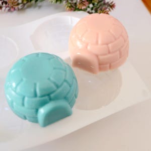 May include: Two igloo-shaped soaps, one light blue and one peach, sit in a white mold. The soaps have a textured brick pattern and a small entrance. The soaps are likely handmade and suitable for a winter-themed gift or decor.