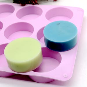 Puede incluir: Primer plano de un molde de silicona rosa con cavidades circulares, que contiene dos jabones redondos. Un jabón es verde claro, el otro es azul. Los jabones son hechos a mano y tienen una superficie lisa. El molde está diseñado para hacer jabón.