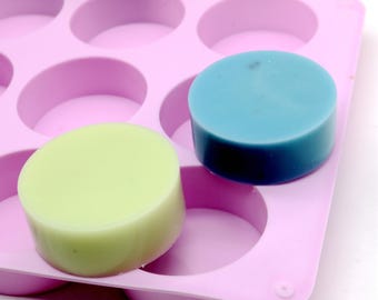 Puede incluir: Primer plano de un molde de silicona rosa con cavidades circulares, que contiene dos jabones redondos. Un jabón es verde claro, el otro es azul. Los jabones son hechos a mano y tienen una superficie lisa. El molde está diseñado para hacer jabón.