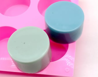 May include: Two round bars of soap, one light blue and one light green, are sitting in a pink silicone mold. The mold has six circular cavities.