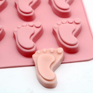 May include: Pink silicone mold with six baby footprint shapes. One footprint is removed from the mold and is a light pink color.