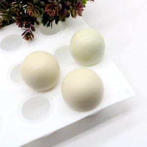 May include: A white silicone mold with several round cavities, holding three pale yellow spheres. The spheres are smooth and evenly colored. The mold is on a white surface, with a few decorative plants in the background.