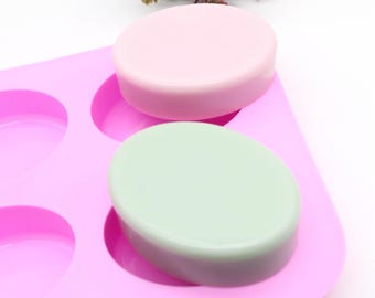 May include: A pink silicone soap mold with oval-shaped cavities, holding two oval soaps. One soap is light pink, the other is light green. The mold is on a white background with some greenery.