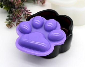Può includere: Uno stampo in silicone a forma di impronta di zampa di colore viola con cinque incavi per realizzare sapone o artigianato in resina. Lo stampo è posto sopra un contenitore nero a forma di impronta di zampa.