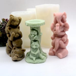 Op de afbeelding: Drie decoratieve kaarsenhouders gemaakt van lichtbruin, roze en groen materiaal. De bruine houder heeft een stapel van drie wasberen, de roze houder heeft een stapel van drie olifanten en de groene houder heeft een vogelbad op een boomstam met drie wasberen.