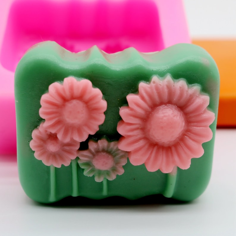 Puede incluir: Una pastilla de jab&oacute;n verde con tres flores de margarita rosas en la parte superior. La pastilla de jab&oacute;n est&aacute; en un molde de silicona.