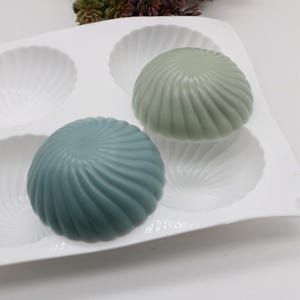 May include: A white silicone mold with six round depressions, two of which are filled with teal and green soap. The soap is shaped like a seashell with a swirl pattern.