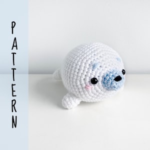 May include: White crocheted seal amigurumi toy with blue and pink details. The seal has a small, round body with a cute face and black eyes.